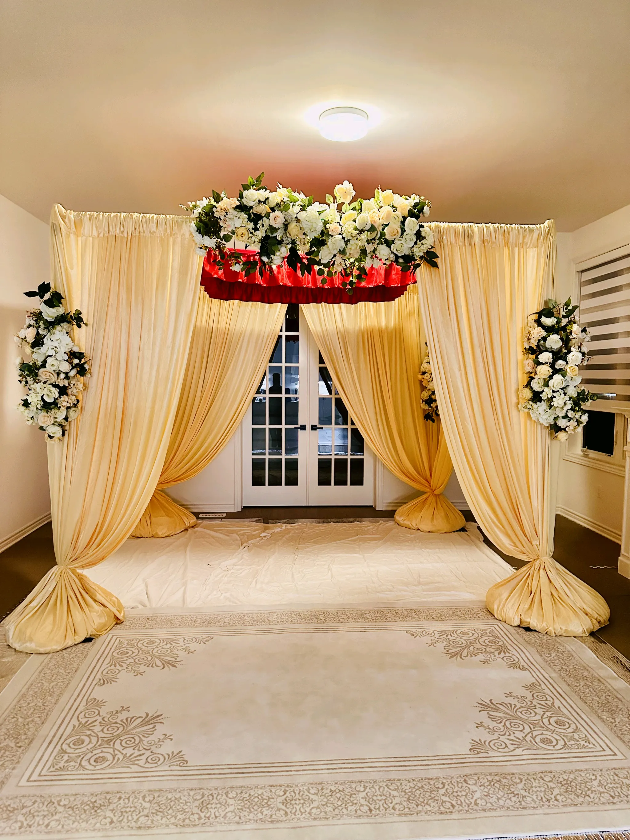 Traditional Indian wedding mandap featuring gold satin drapery, lush white and cream floral arrangements, a red fabric accent, and an ornate cream rug in an indoor event setting.