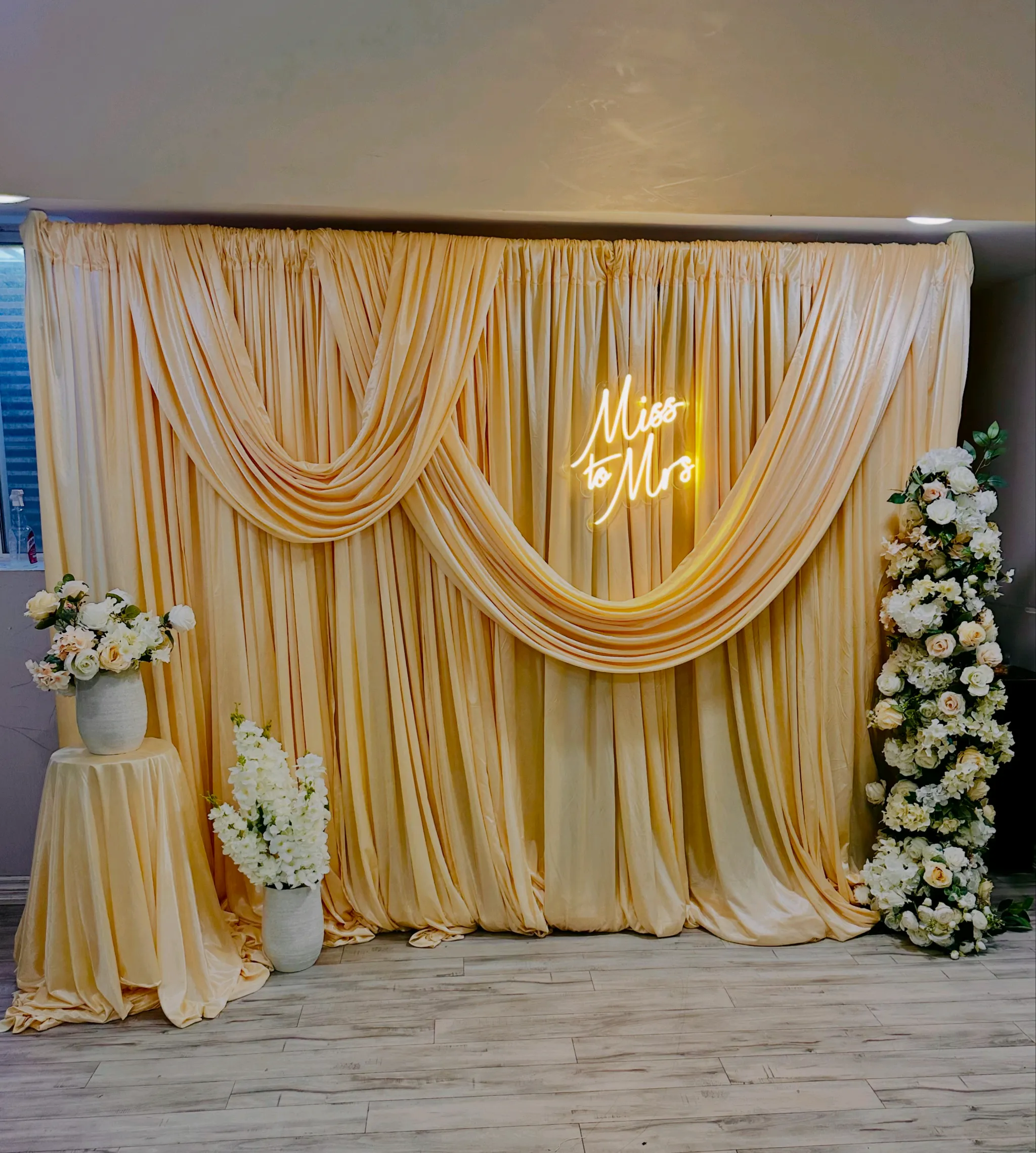 Elegant champagne gold satin fabric backdrop for a bridal shower featuring a 'Miss to Mrs' neon sign, white floral arrangements, and a decorative floral pillar in a modern indoor event setting.