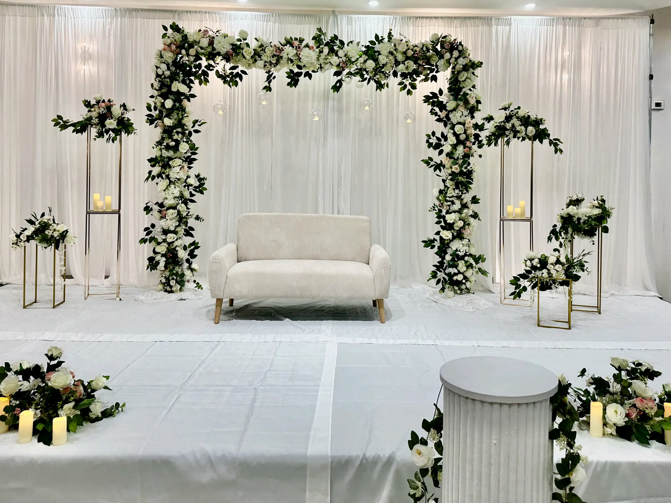Elegant white wedding event stage decor featuring a cream loveseat, a large white and blush pink floral arch, gold metal flower stands, and sheer white curtains for a romantic atmosphere.